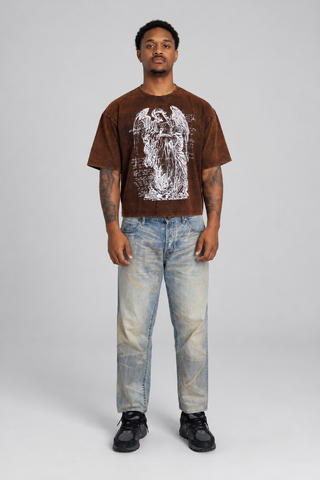 Front view of a model wearing the brown washed Guardian Cropped T-Shirt, showing the angel graphic, boxy fit, cropped hem, faded jeans, and black sneakers.
