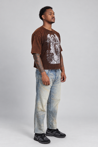 Left three-quarter view of a model wearing the brown washed Guardian Cropped T-Shirt, showing the angel graphic, sleeve volume, drape, cropped fit, faded jeans, and black sneakers.