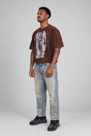 Right three-quarter view of a model wearing the brown washed Guardian Cropped T-Shirt, showing the angel graphic, sleeve volume, drape, cropped fit, faded jeans, and black sneakers.