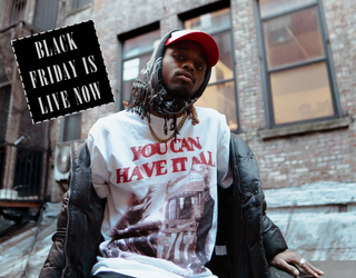 Person wearing a white t-shirt with text and graphics, sitting on a ledge with a brick building in the background.