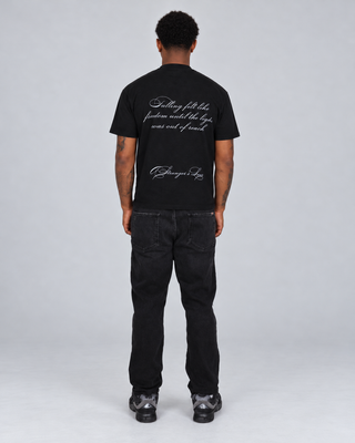 Full-body back view of a model wearing the black Fallen T-Shirt, showing the script quote and A Stranger's Love logo, with black jeans and dark sneakers.